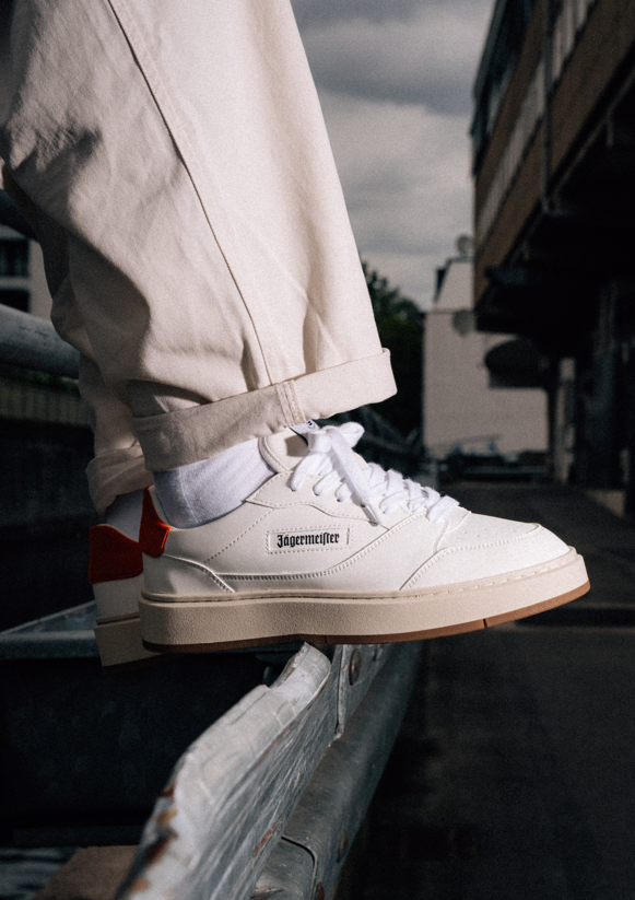 A person wearing off-white Jägermeister sneakers with white socks and beige pants, sitting on a metal railing in an outdoor urban setting.
