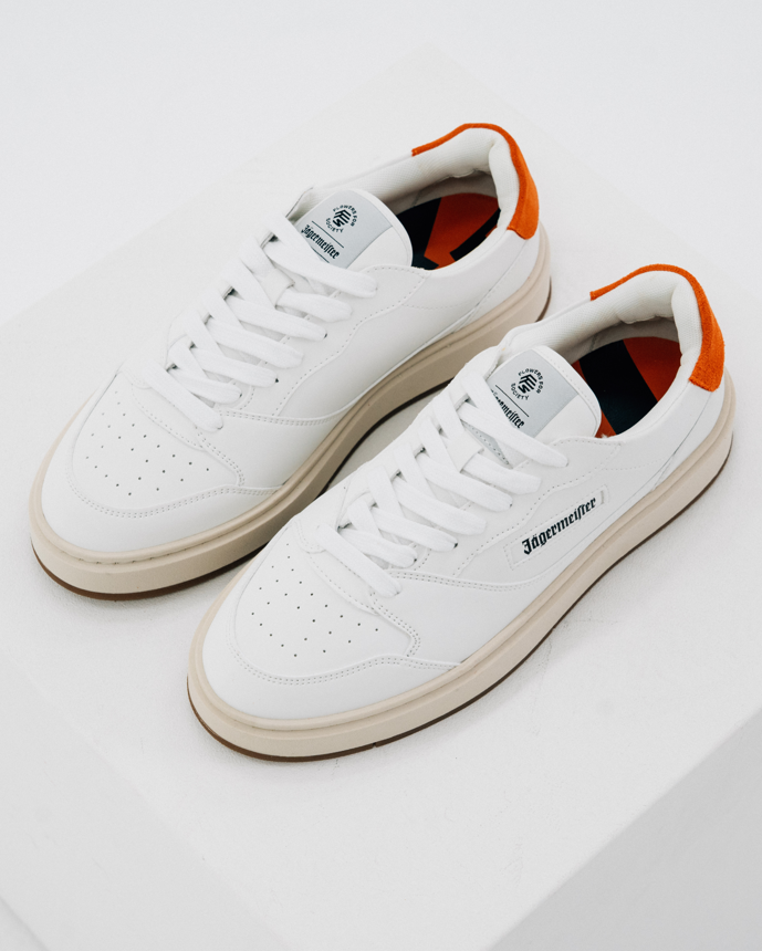 A pair of white Jägermeister sneakers with orange heel accents and beige midsoles, photographed on a clean white studio background. The brand name 'Jägermeister' is visible on the side of the shoe.