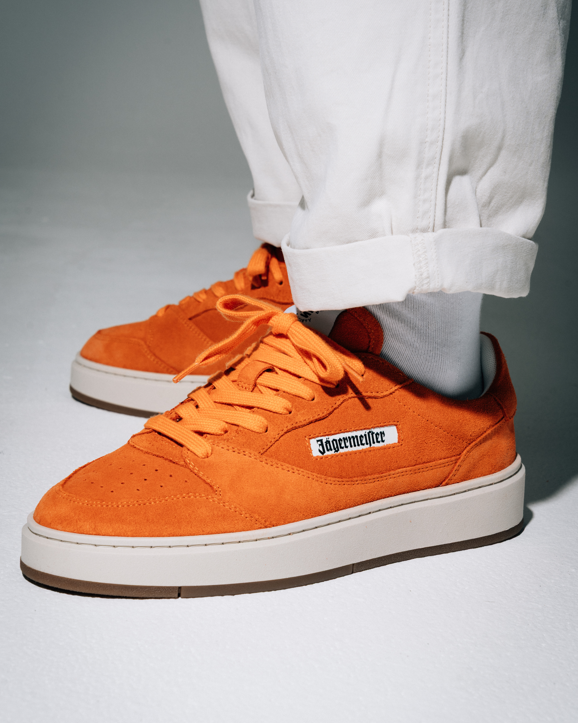 A person wearing bright orange Jägermeister sneakers with white cuffed pants, shown from the knees down on a light gray studio background. The shoe has a white side label reading 'Jägermeister' and a white midsole with a gum outsole.