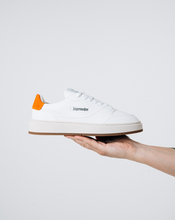 A white Jägermeister sneaker with a bright orange heel tab and a black 'Jägermeister' wordmark on the side, held up by a person's hand against a light background.