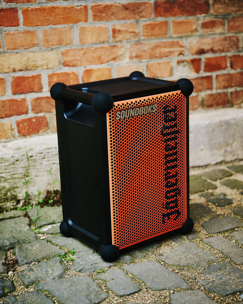 Boxă portabilă Soundboks portocalie și neagră, cu branding Jägermeister, lângă un zid de cărămidă