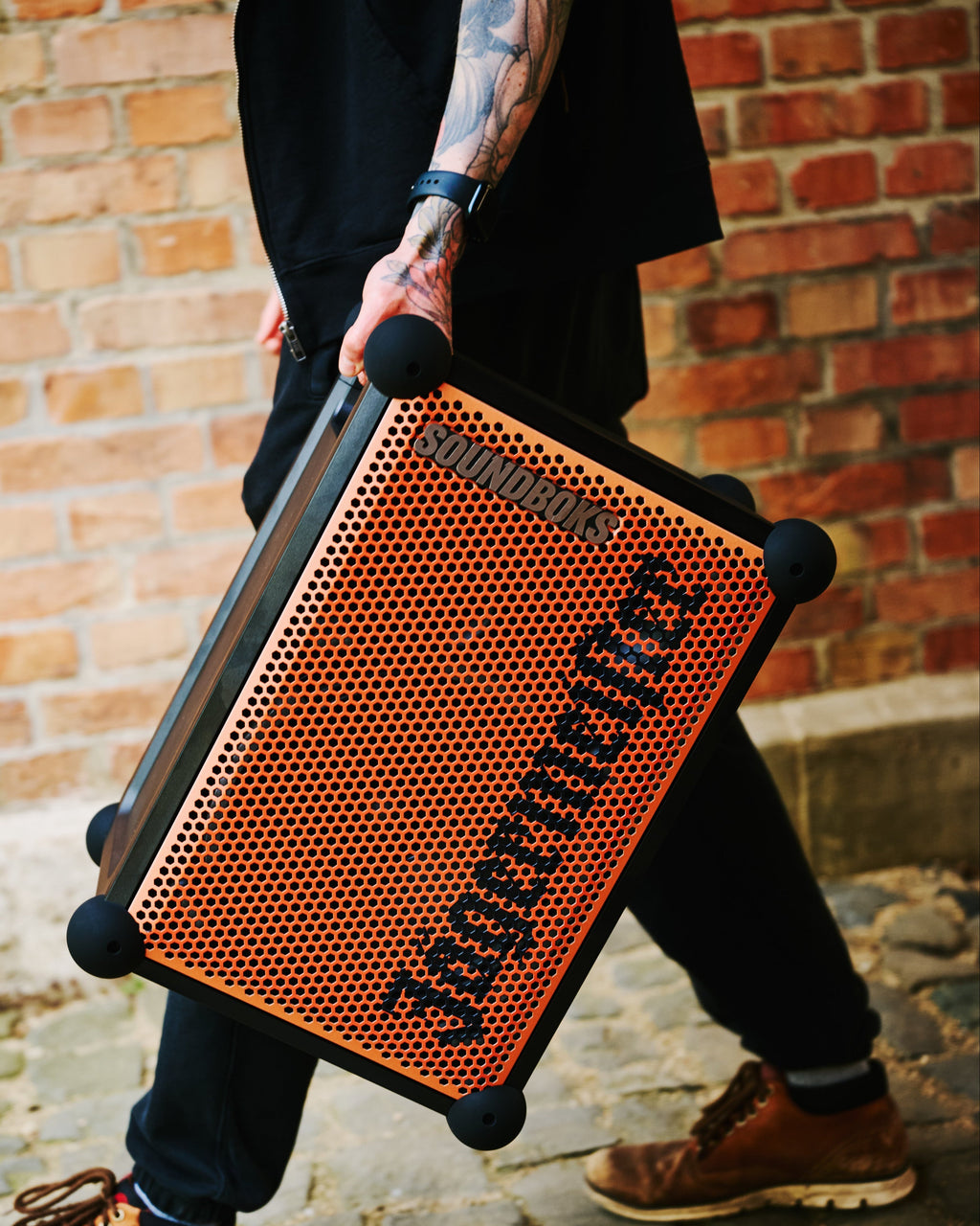 Persoană care ține o boxă portabilă Soundboks portocalie și neagră, cu branding Jägermeister, în fața unui zid de cărămidă.