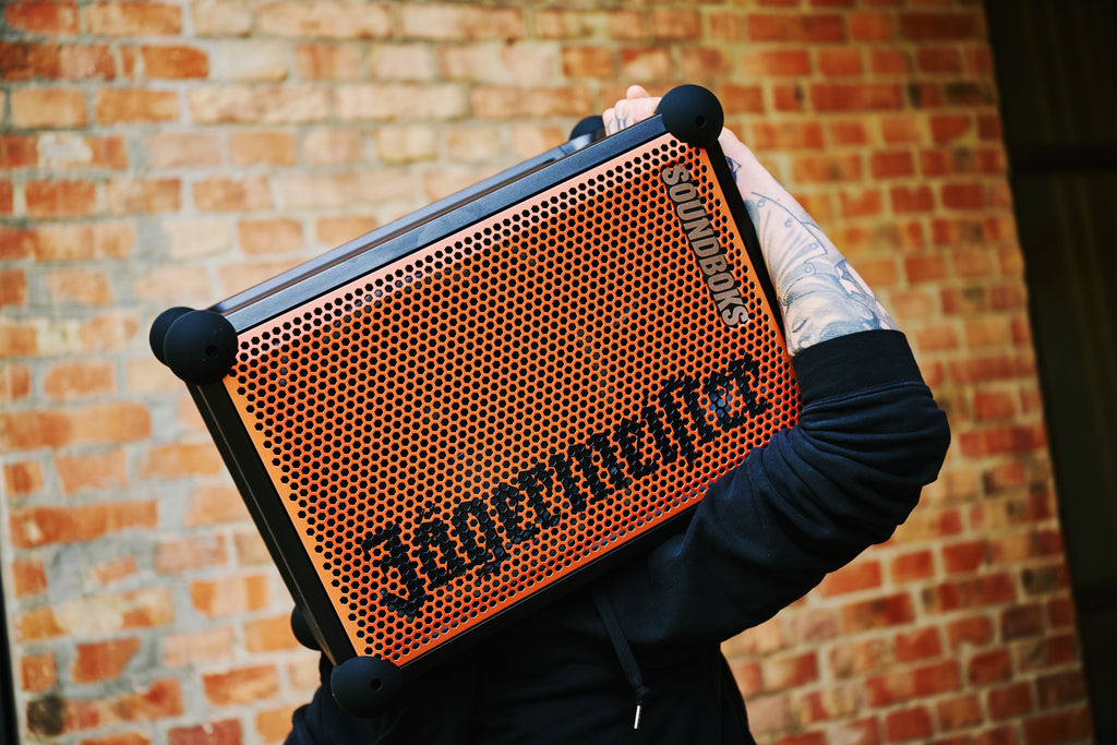 Persoană care ține o boxă portabilă Soundboks portocalie și neagră, cu branding Jägermeister, în fața unui zid de cărămidă.