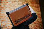 Persoană care ține o boxă portabilă Soundboks portocalie și neagră, cu branding Jägermeister, în fața unui zid de cărămidă.