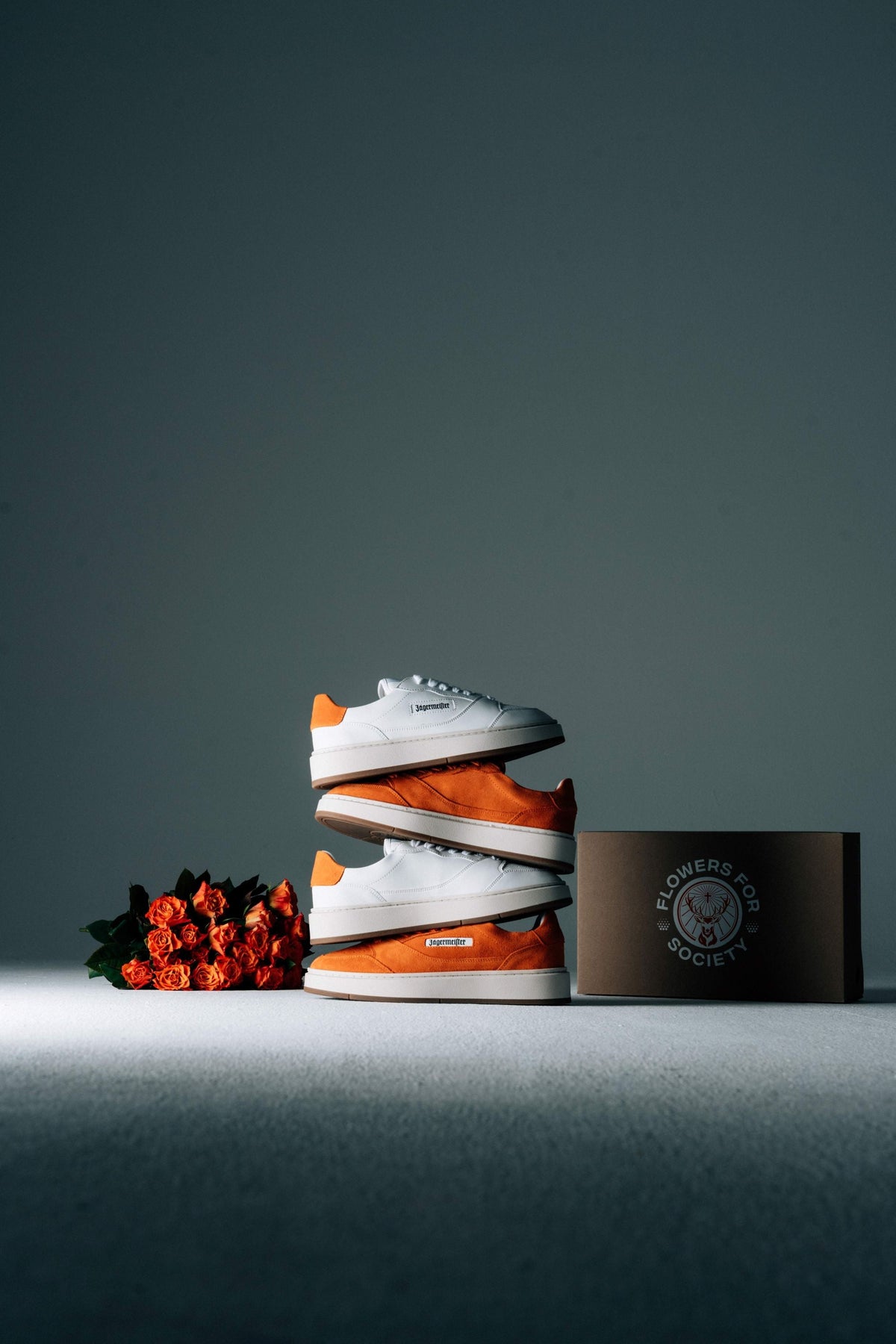 Two pair of sneakers displayed next to a dark brown packaging box with the Jägermeister and Flowers for Society logo and a small bouquet of red-orange flowers on a dark gray surface.