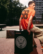 Un bărbat într-un tricou de fotbal Jägermeister portocaliu și pantaloni bej stă pe o boxă Soundboks neagră cu logo-ul Jägermeister