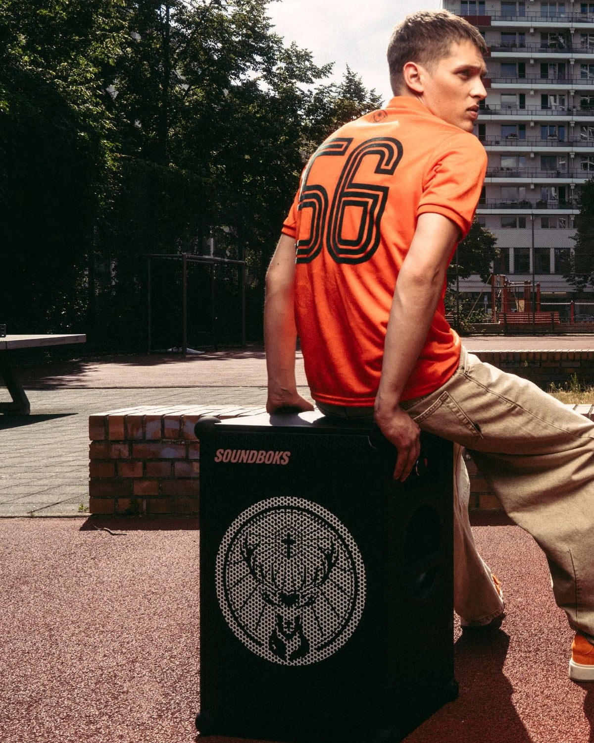 Un bărbat într-un tricou de fotbal Jägermeister portocaliu și pantaloni bej stă pe o boxă Soundboks neagră cu logo-ul Jägermeister