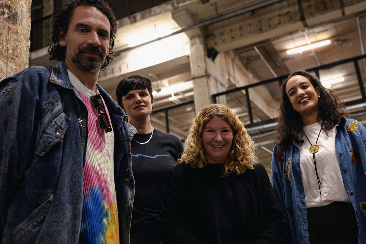Four people standing together in an industrial loft space with exposed ceiling beams and lighting fixtures. The group includes a man on the left in a denim jacket over a colorful tie-dye shirt, a woman behind him in a dark top, a woman in the center foreground in a black top, and a woman on the right in a blue jacket over a white top.