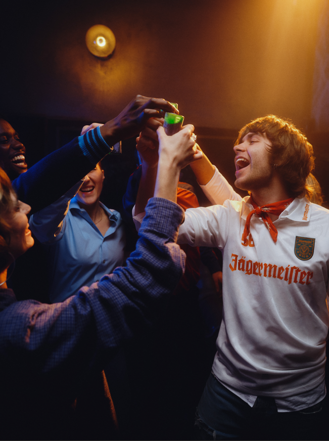 Un grup de persoane sărbătorind alături de o persoană care poartă un tricou Jägermeister, într-o cameră slab iluminată.